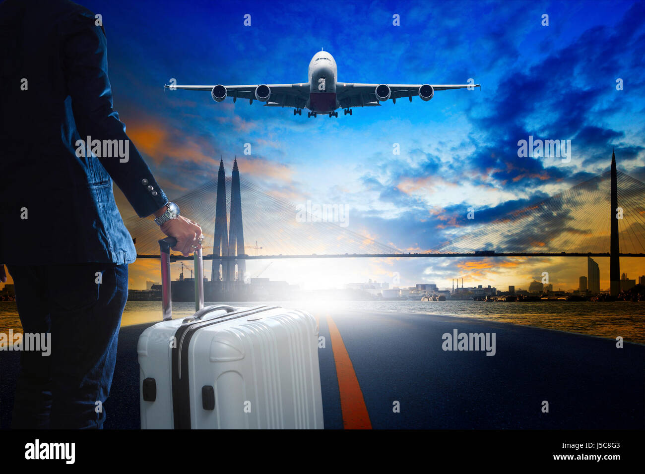 young business man standing with luggage on urban airport runway and jet plane flying above against beautiful urban scen behind Stock Photo