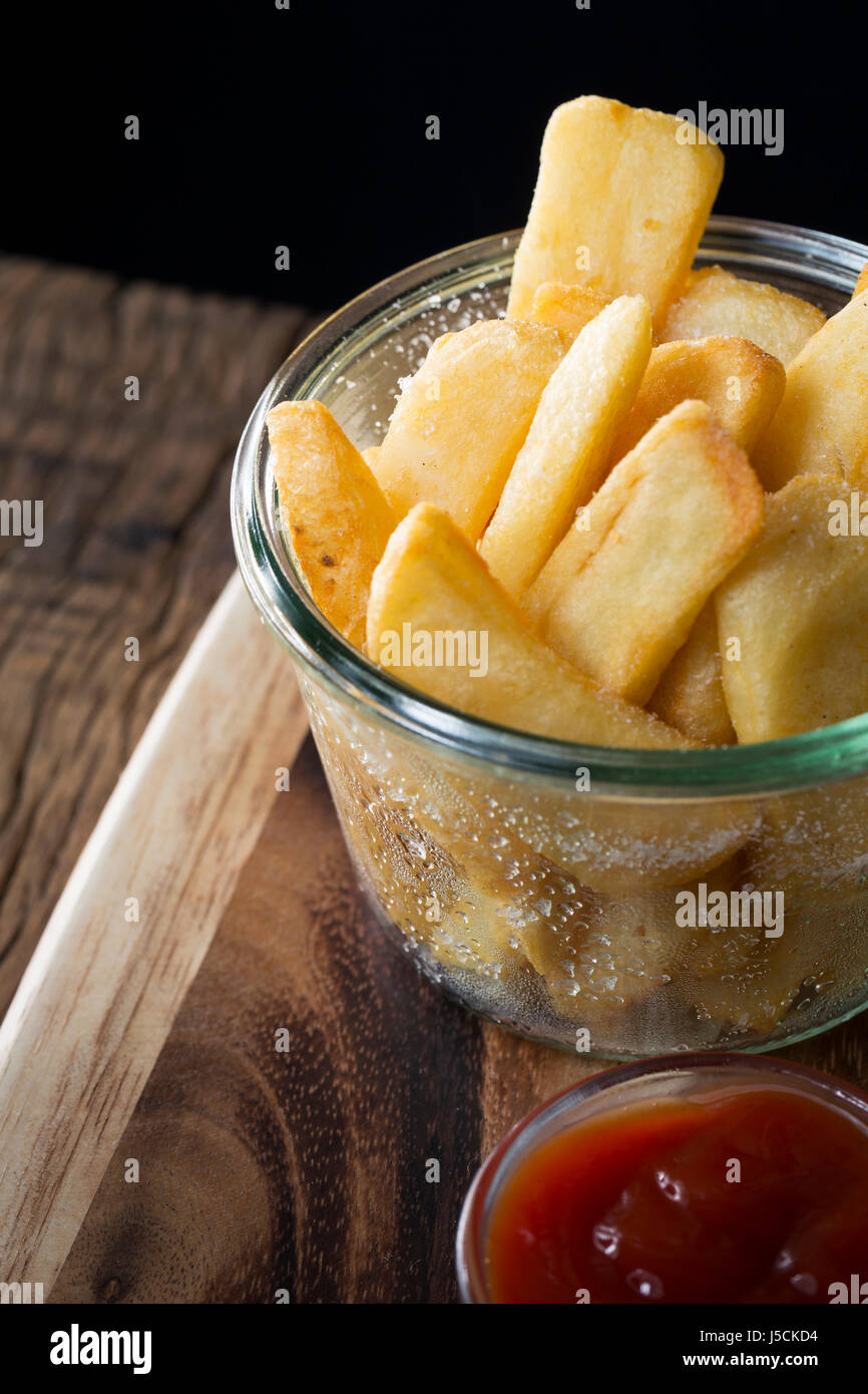 Close up of fresh french fries or hot potato chips served in a glass bowl. The food is sitting on a rustic wooden background. Stock Photo