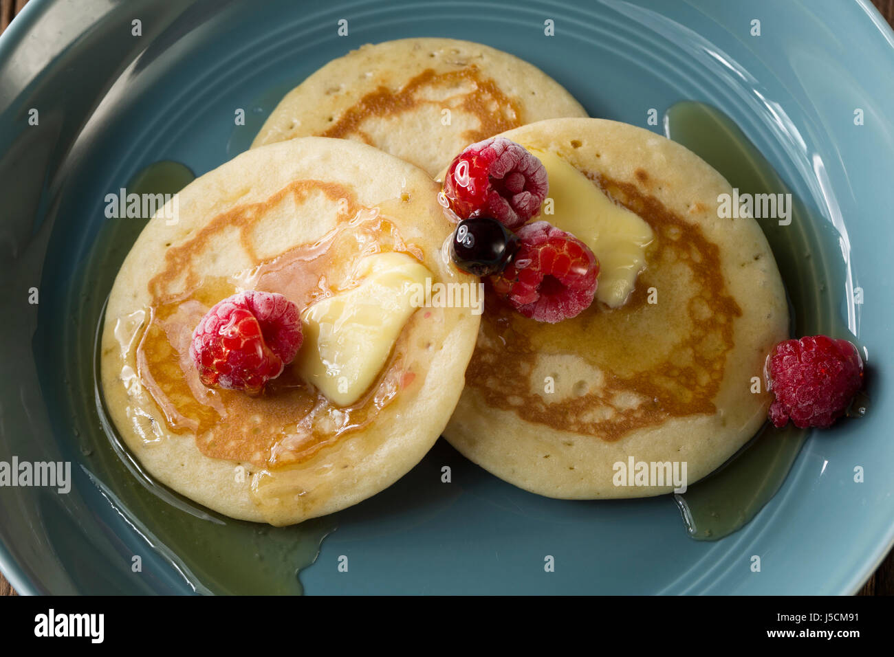 Three fresh pancakes, drizzled with syrup and Berries in a blue bowl, sitting on a rustic wooden table. Stock Photo