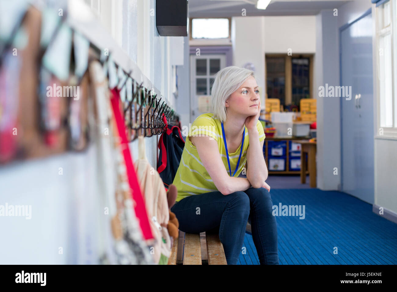 Young, female teacher sitting on a bench in the cloakroom of school. She is looking pensively out of the window. Stock Photo