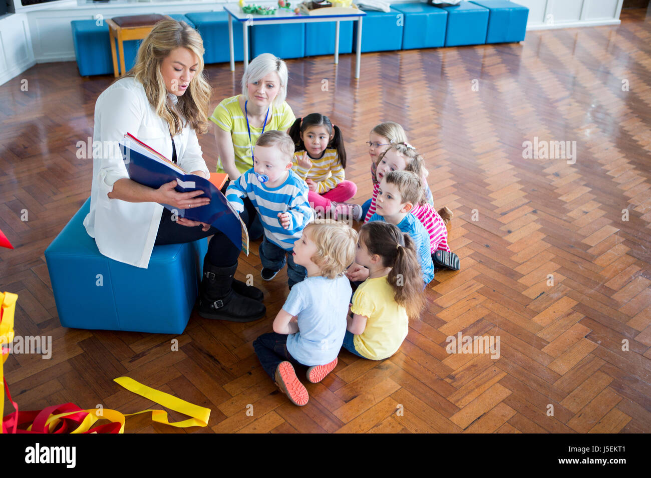 Small group of nursery students sitting around a teacher in their school hall. She is reading a book to them. Stock Photo
