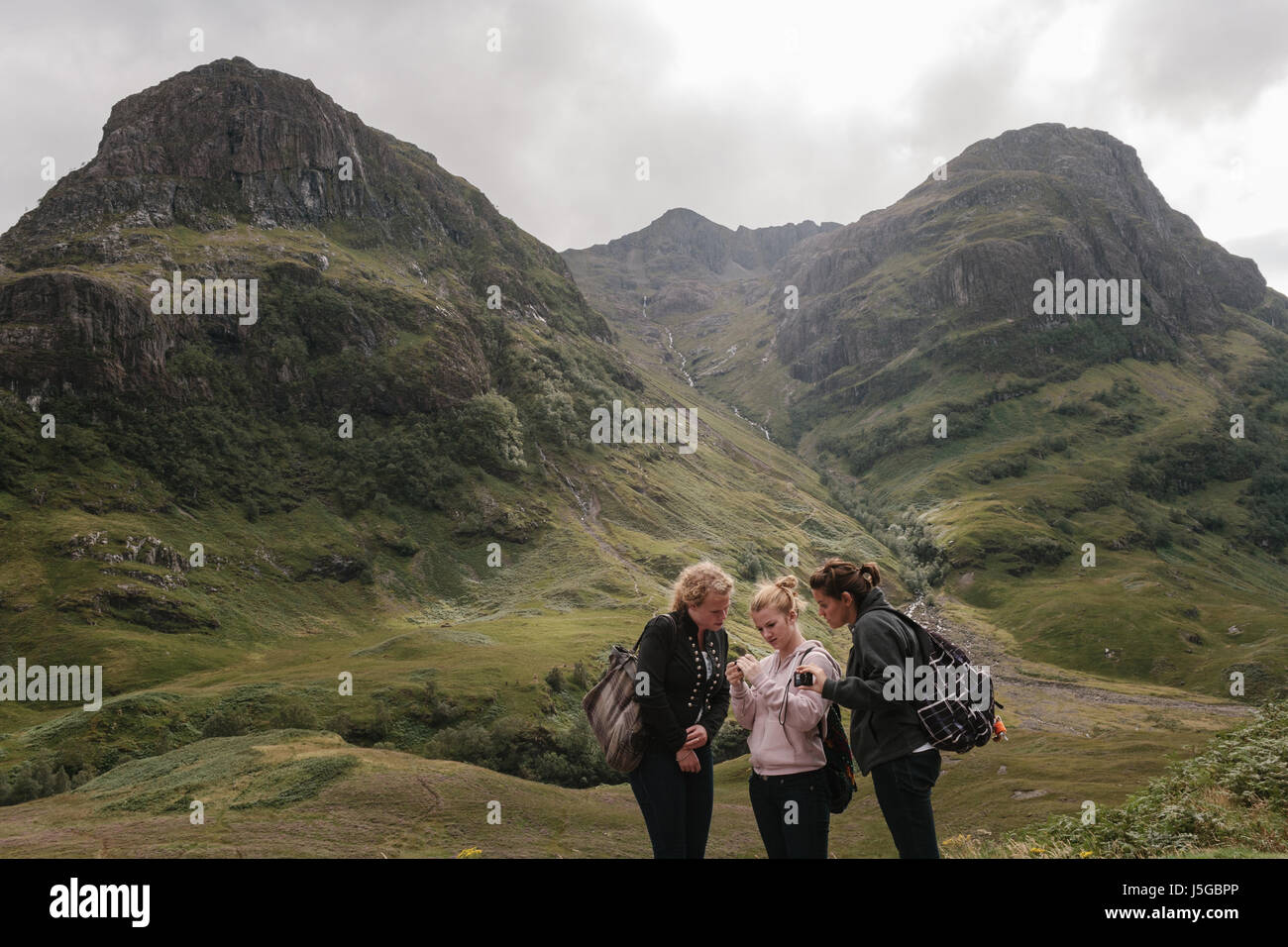 Three attractive female tourists looking at photographs on their smart phones with the mountains behind them in Glencoe, Scotland. Stock Photo