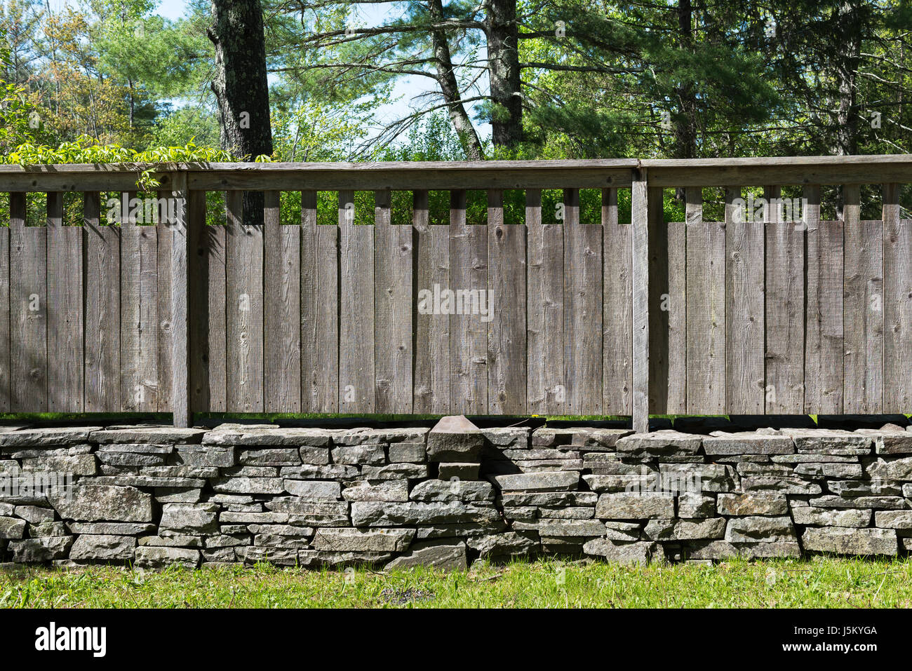 Section of Stacked Stone Wall with Grey Distressed Wood Fence in Country Setting . Grass in foreground and Trees in Background. Stock Photo