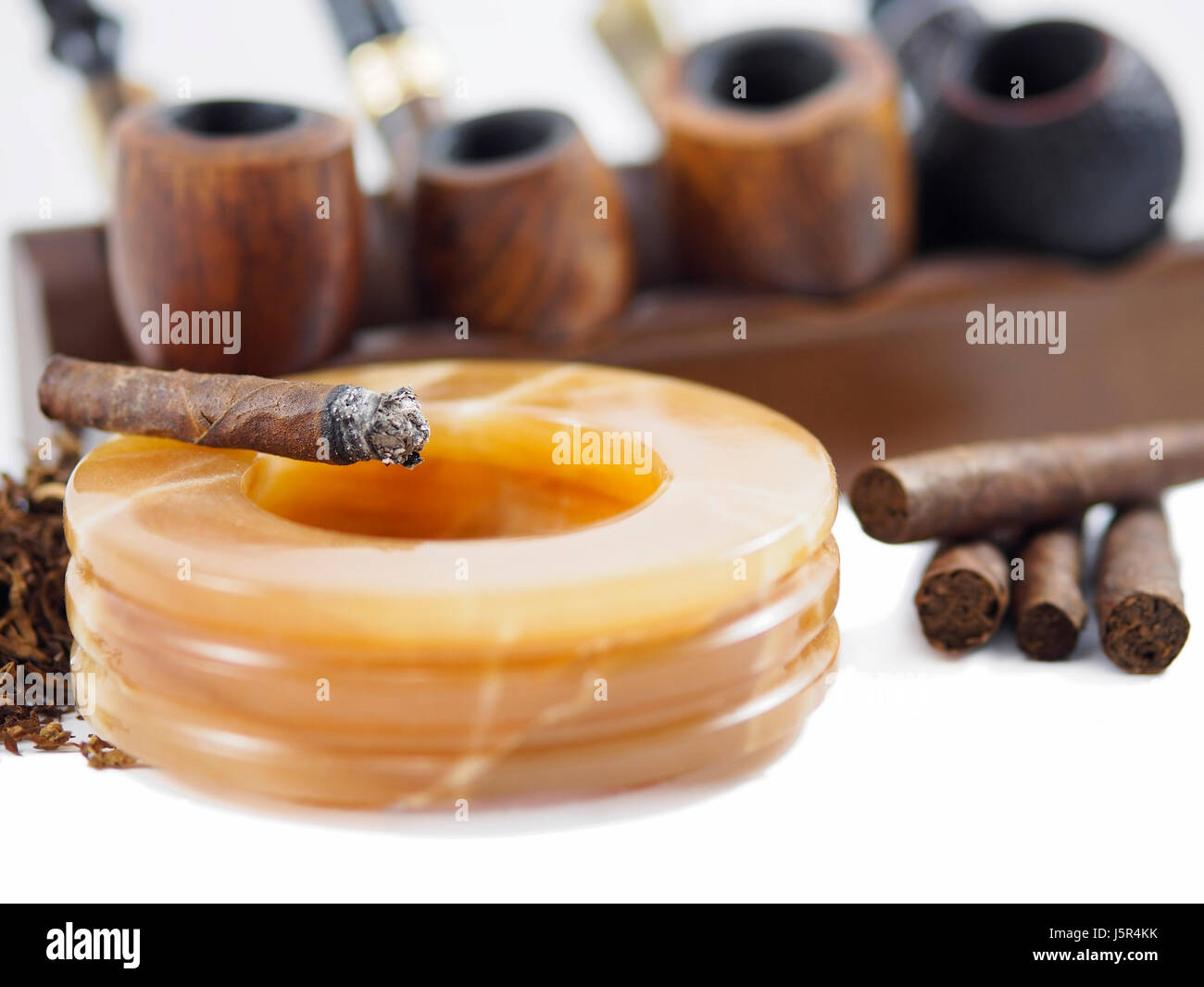Burning Toscano cigar on an alabaster ashtray. On the background, four Italian pipes. Stock Photo