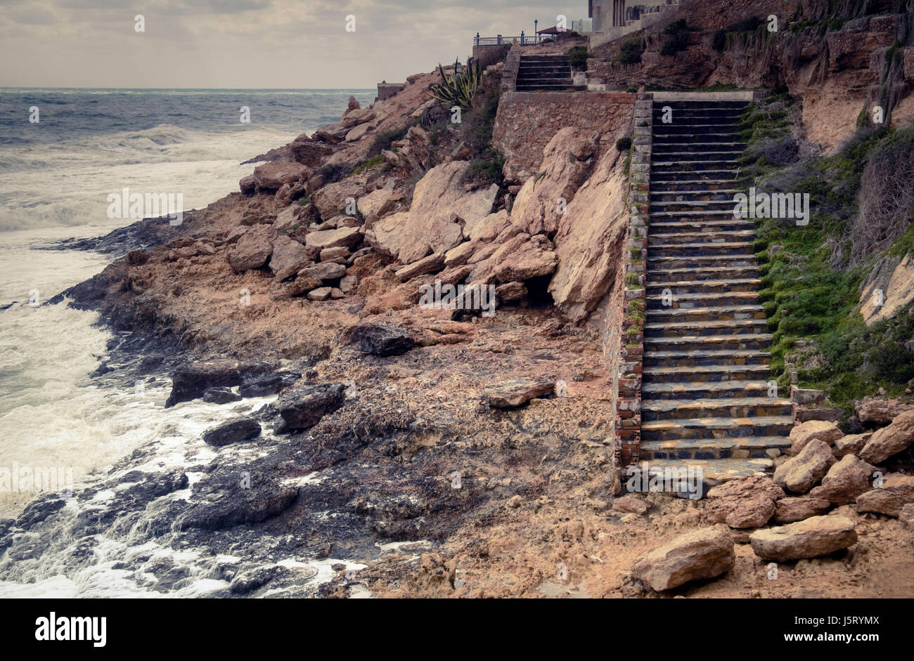 An overcast image of a weathered stairway built into cliff and rocks along the Spanish coastline. Stock Photo