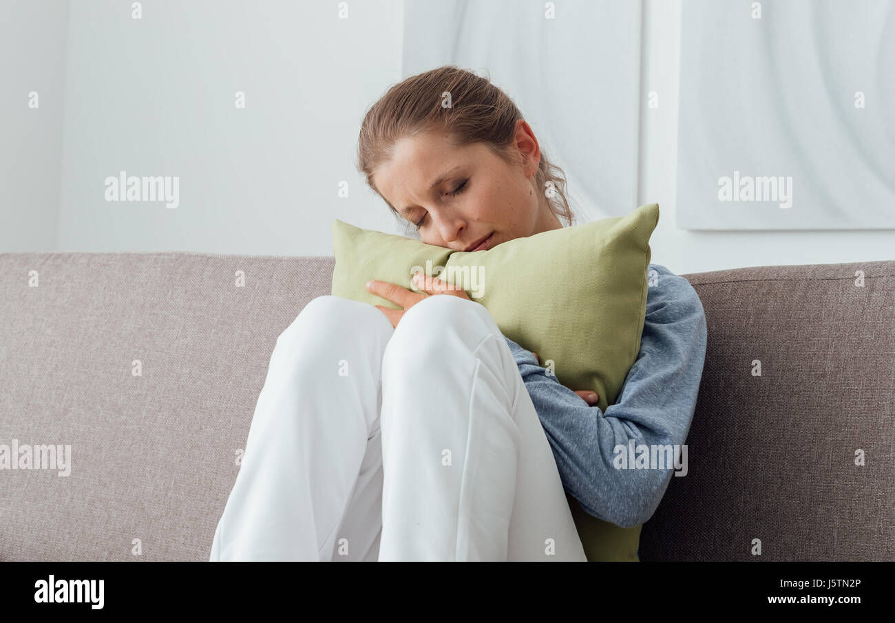 Sad depressed woman at home, she is sitting on the couch and hugging a ...