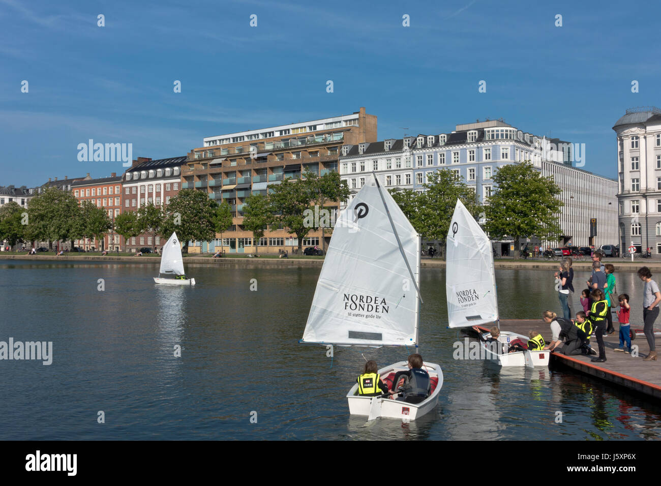 Junior sailors in optimist dinghies coached on the Peblinge Lake in central Copenhagen on a sunny afternoon in early summer. Parents on the jetty. Stock Photo