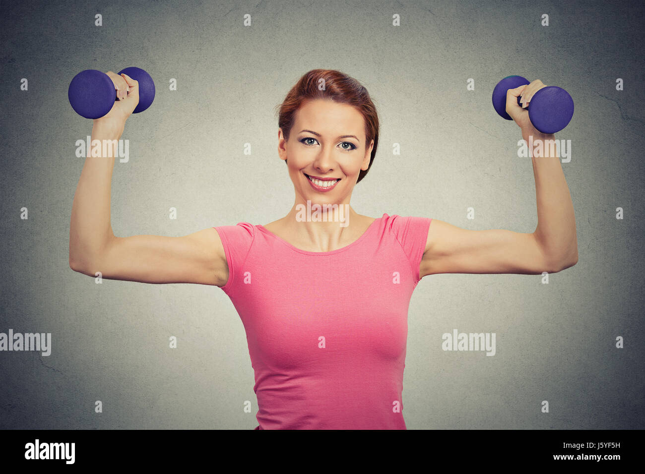 Closeup portrait beautiful fit healthy model woman flexing muscles lifting dumbbells confident showing her strength isolated grey background. Positive Stock Photo