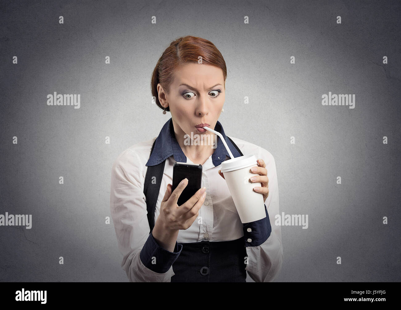 Closeup portrait surprised business woman reading breaking news on smart phone holding mobile drinking soda coffee isolated grey wall background. Huma Stock Photo
