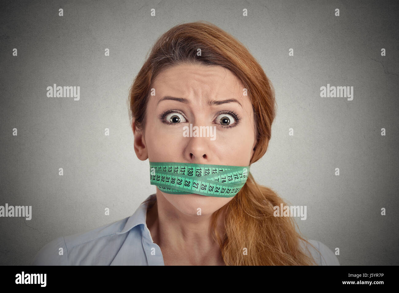 Portrait unhappy young woman with green measuring tape covering mouth isolated grey wall background. Dieting lifestyle concept Stock Photo