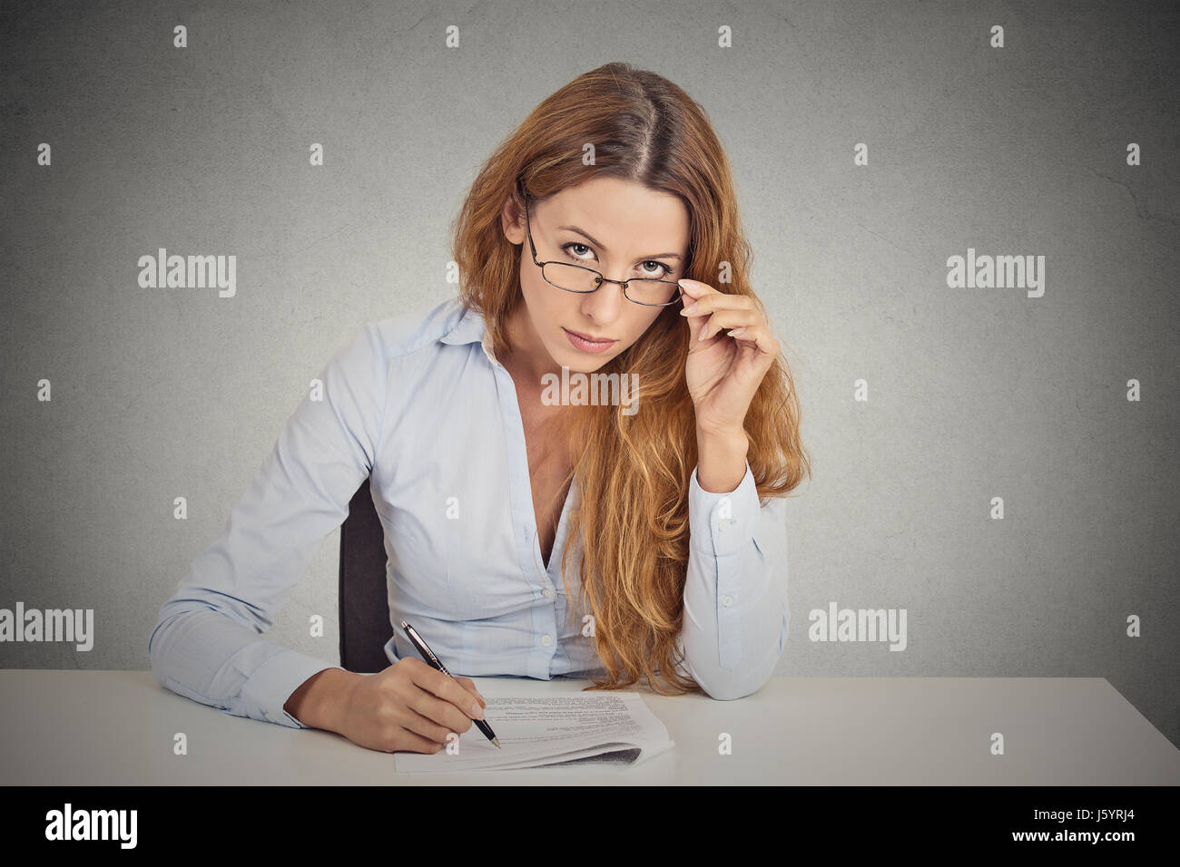 Curious corporate businesswoman with glasses sitting at desk skeptically looking at you scrutinizing isolated on office grey wall background. Human fa Stock Photo