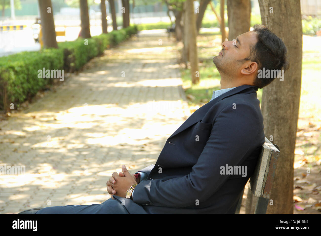 Young Indian corporate man with closed eyes, resting on bench in park Stock Photo