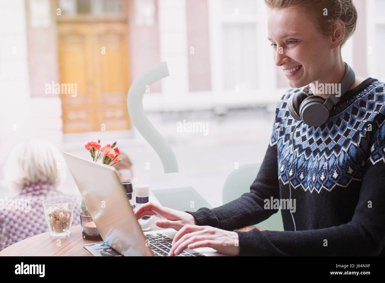 Smiling young woman with headphones using laptop in sidewalk cafe window Stock Photo