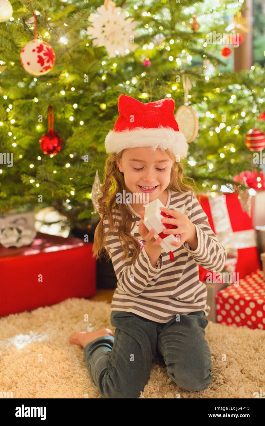 Smiling girl in Santa hat opening gift in front of Christmas tree Stock Photo