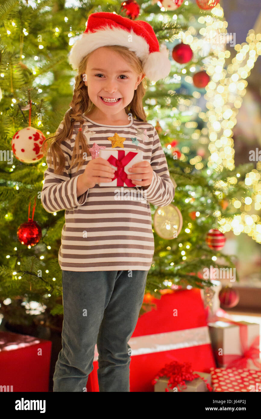 Portrait smiling girl in Santa hat holding gift in front of Christmas tree Stock Photo