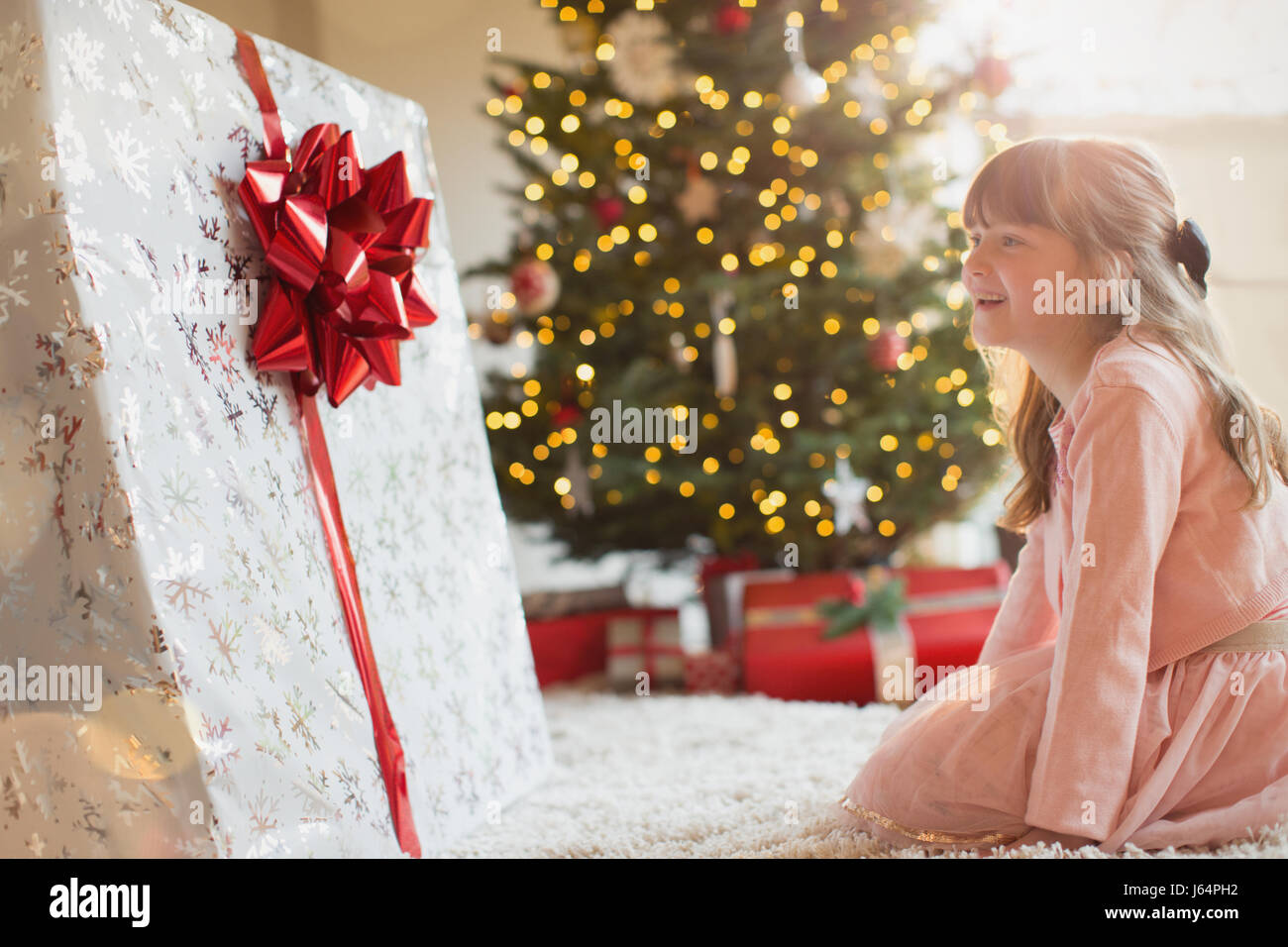 Girl smiling in anticipation at large Christmas gift near Christmas tree Stock Photo