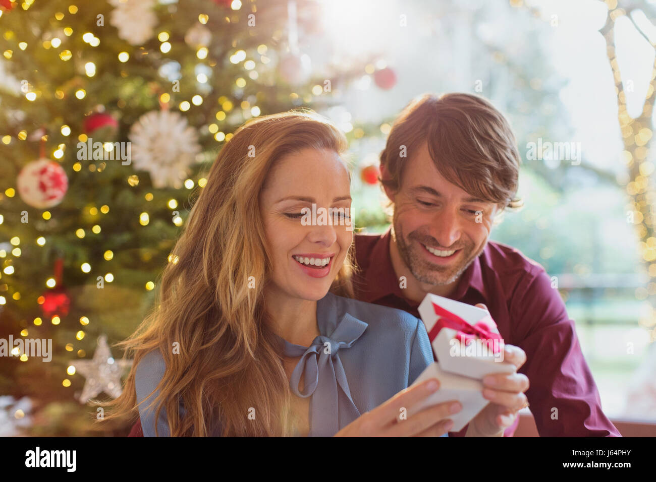 Husband watching wife opening Christmas gift in front of Christmas tree Stock Photo