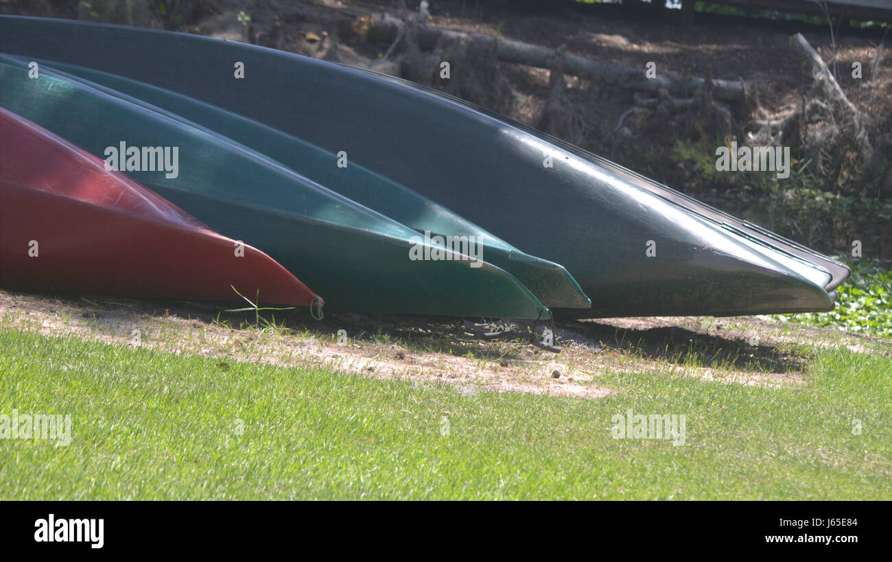 Bottoms of Kayaks and canoes in greens, red, and blues on sandy area surrounded by green grass. Stock Photo