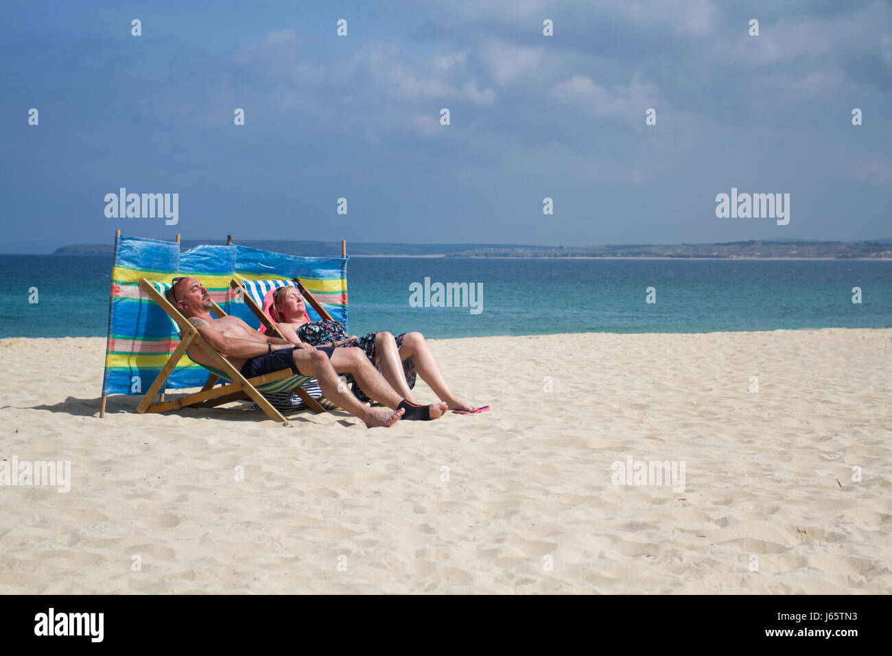 Two British holidaymakers sunbathing in striped deckchairs with a colourful wind break on the sandy beach at Porthminster, Cornwall, UK. Stock Photo
