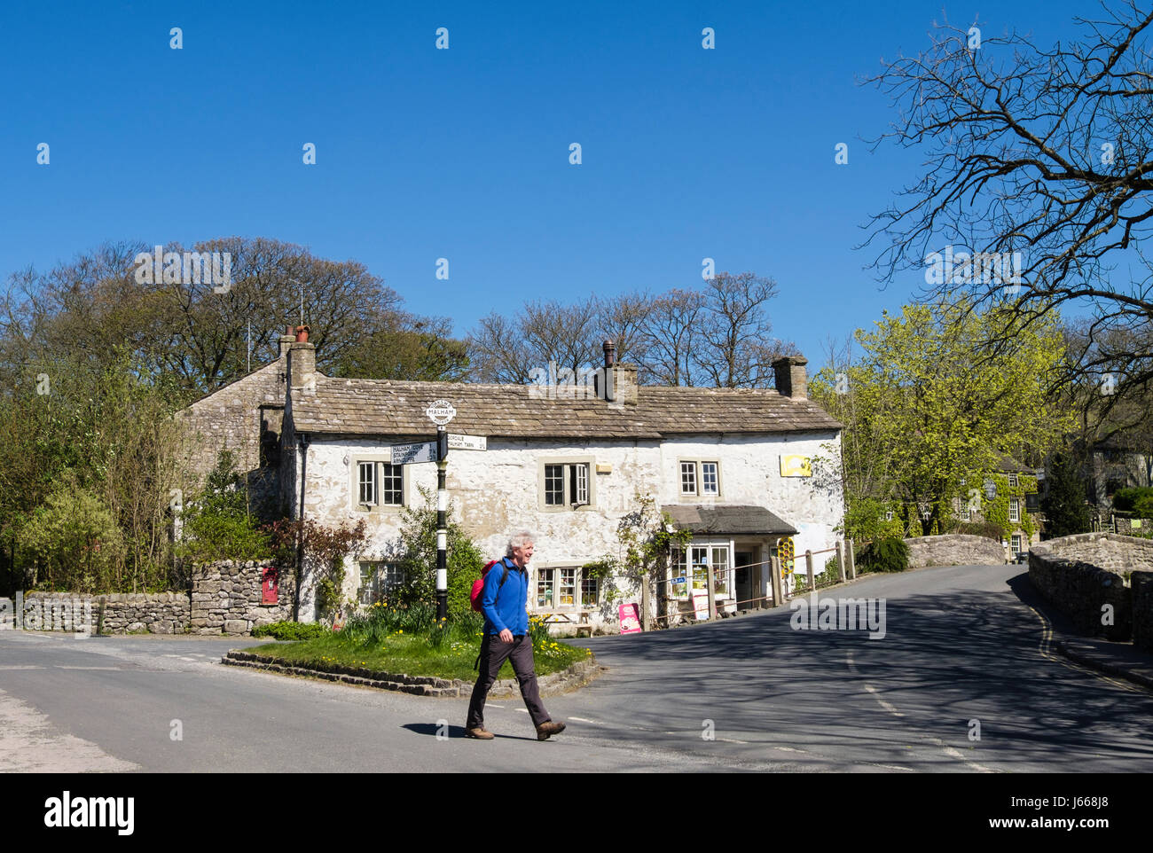 A hiker walking past old shop and signpost at road junction in village centre. Malham Yorkshire Dales National Park North Yorkshire England UK Britain Stock Photo