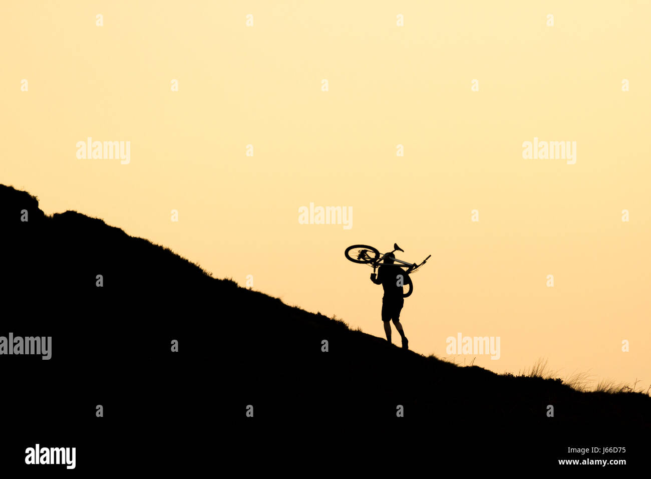 Striking image of a silhouetted mountain biker person carrying his bike uphill, isolated against an orange sky, UK Stock Photo