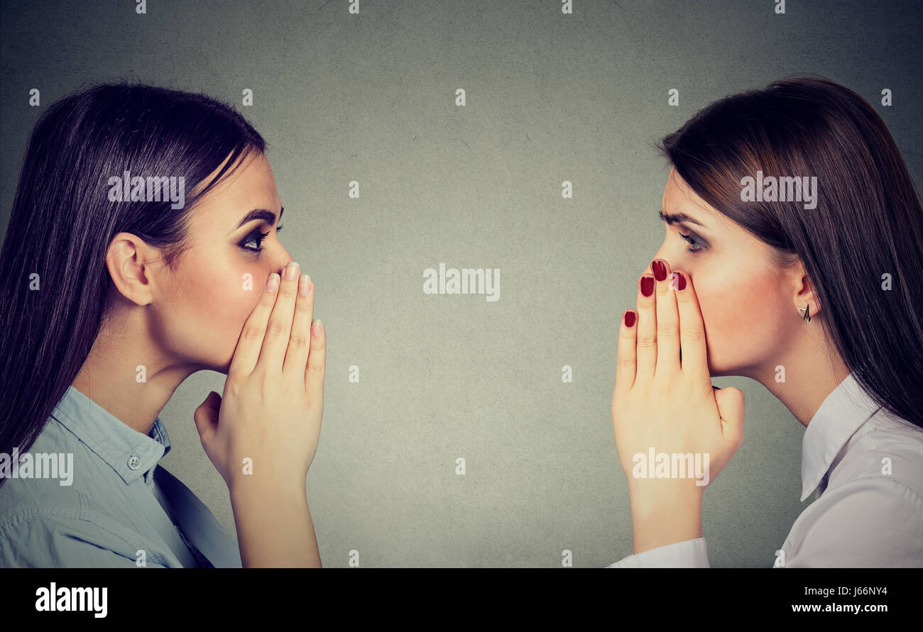 Two women whispering a gossip secret to each other isolated on gray wall background Stock Photo