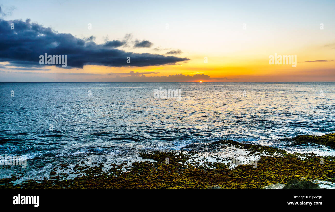 Sunset over the horizon with a few clouds and the rocky shores of the west coast of the tropical Hawaiian island of Oahu Stock Photo