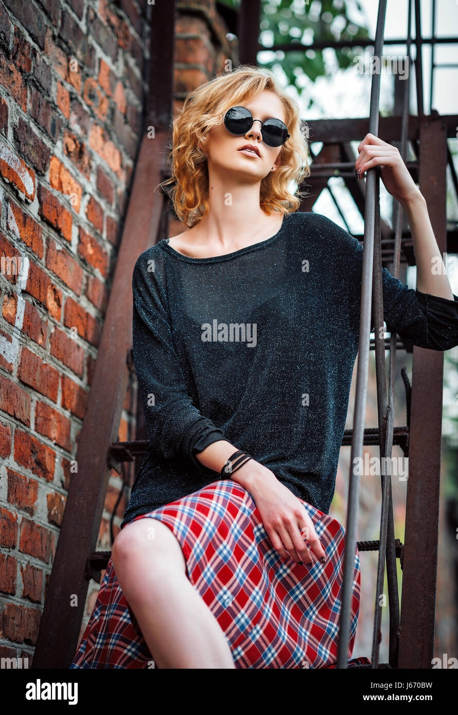 Beautiful young grunge (rock) girl wearing plaid skirt, blouse and glasses sits on the stairway Stock Photo