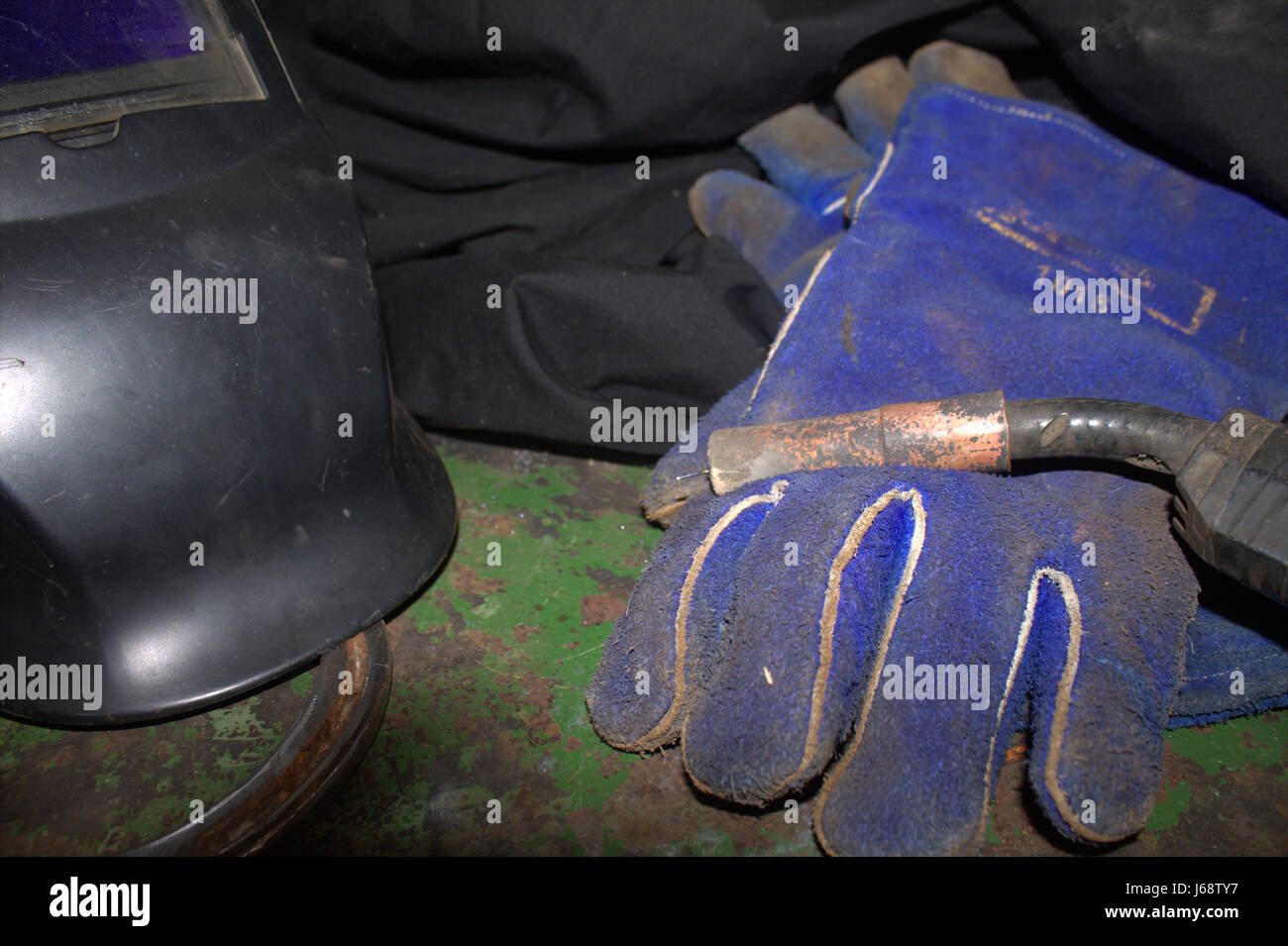 Close up of a welding helmet and welding gun laying on blue welding gloves laying on a green table with peeling paint. Stock Photo