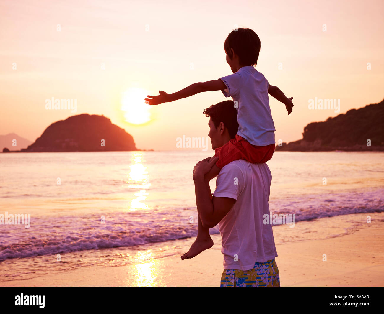 young asian father carrying son on shoulder on beach at sunrise. Stock Photo