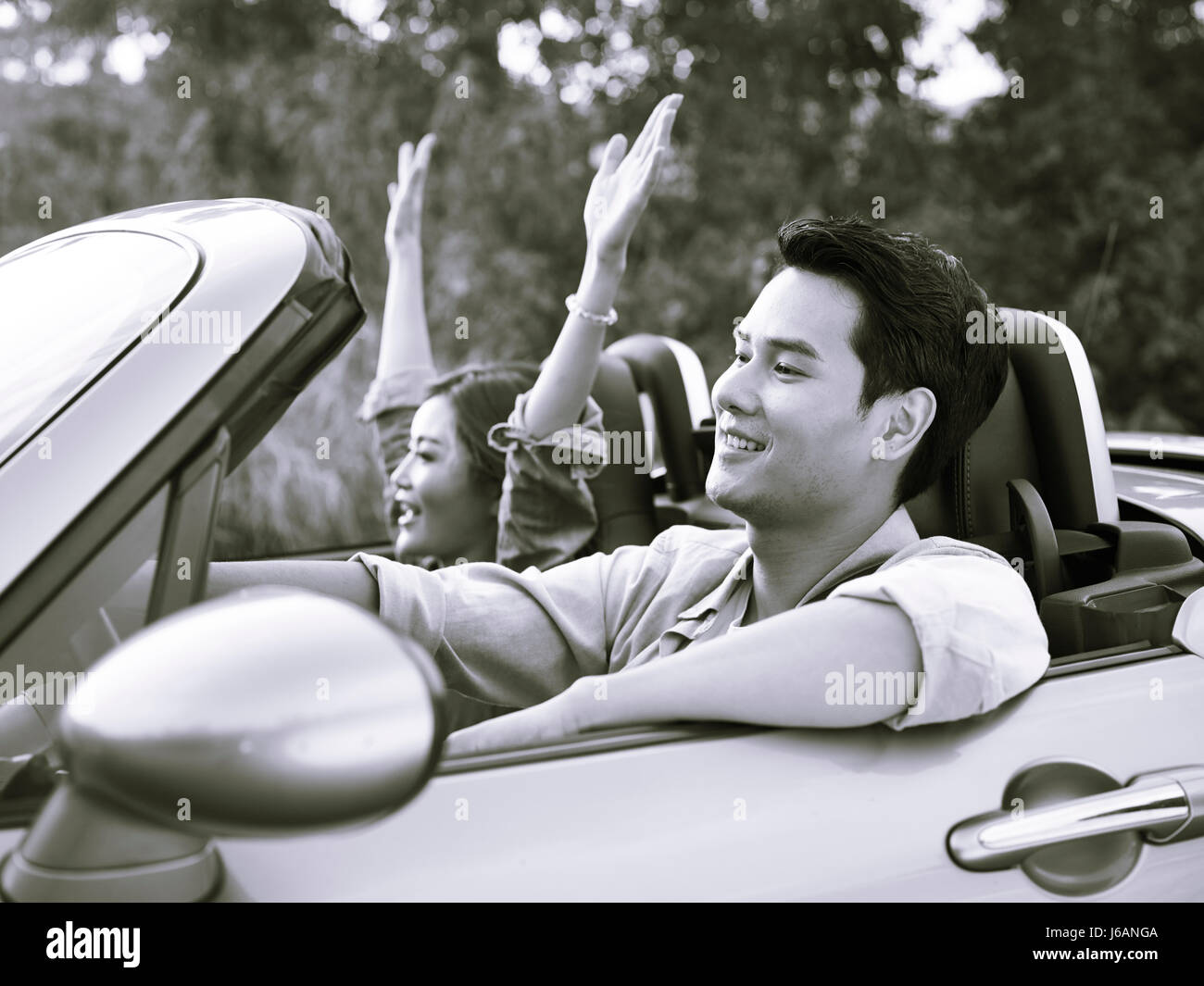 young asian couple riding in a convertible sport car enjoying the cool breeze at sunset, black and white. Stock Photo