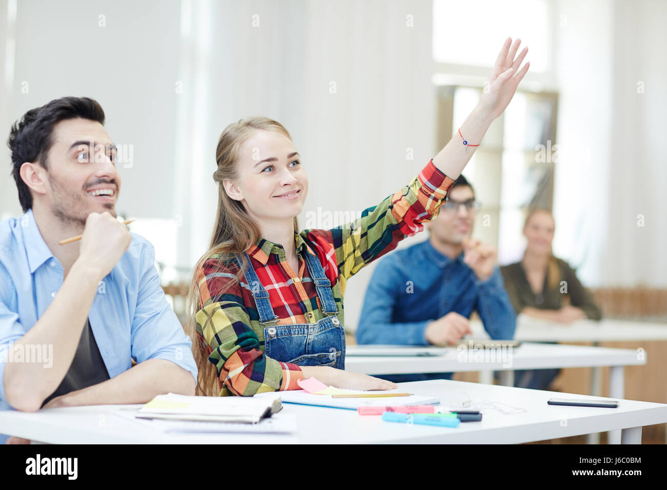 Clever girl raising hand at lesson with groupmate near by Stock Photo