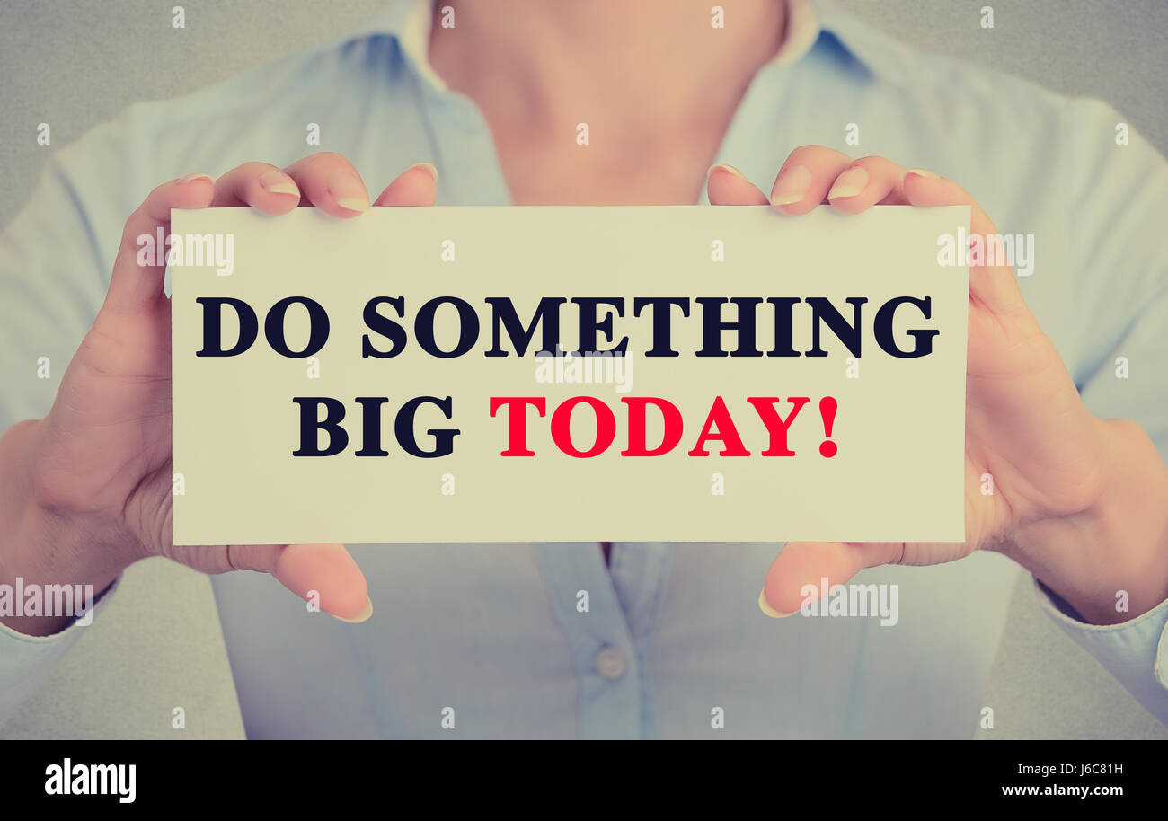 Businesswoman hands holding white card sign with do something big today text message isolated on grey wall office background. Retro instagram style im Stock Photo