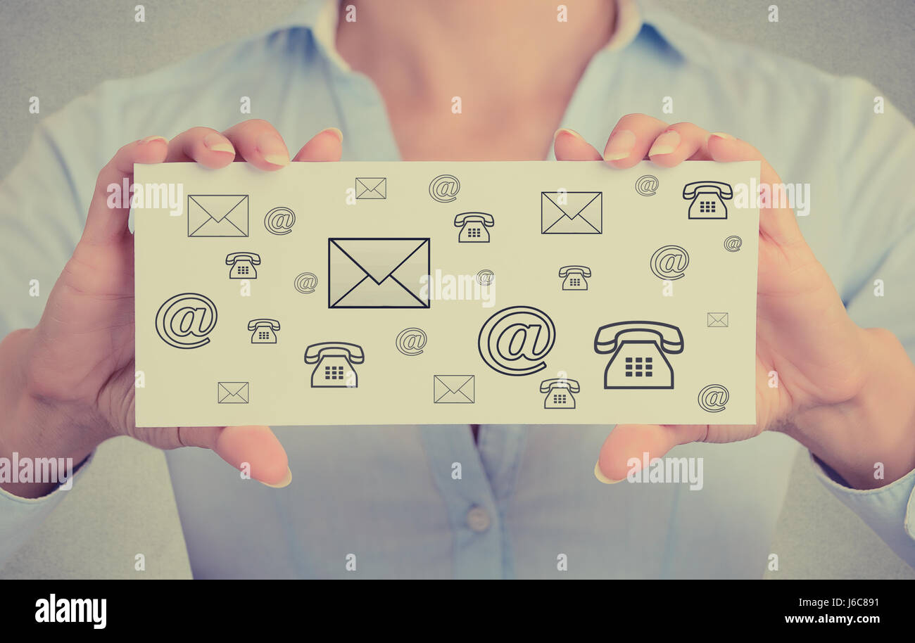 Businesswoman hands holding white card sign with variety of different contact icons for mail, email, web and telephone message isolated on grey wall o Stock Photo
