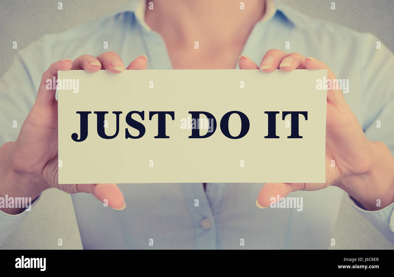 Closeup businesswoman hands holding white card sign with just do it text message isolated on grey wall office background. Retro instagram style image Stock Photo