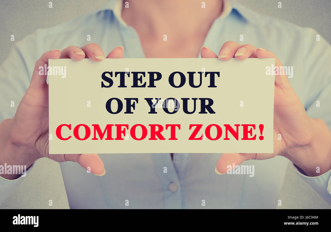 Businesswoman hands holding white card sign with step out of your comfort zone text message isolated on grey wall office background. Retro instagram s Stock Photo