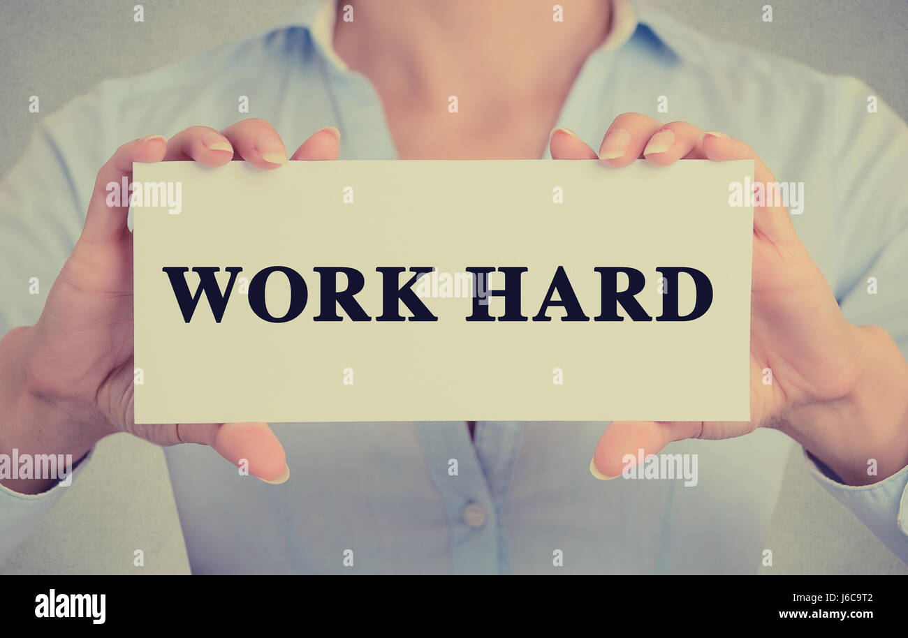 Closeup businesswoman hands holding white card sign with work hard text message isolated on grey wall office background. Retro instagram style image Stock Photo