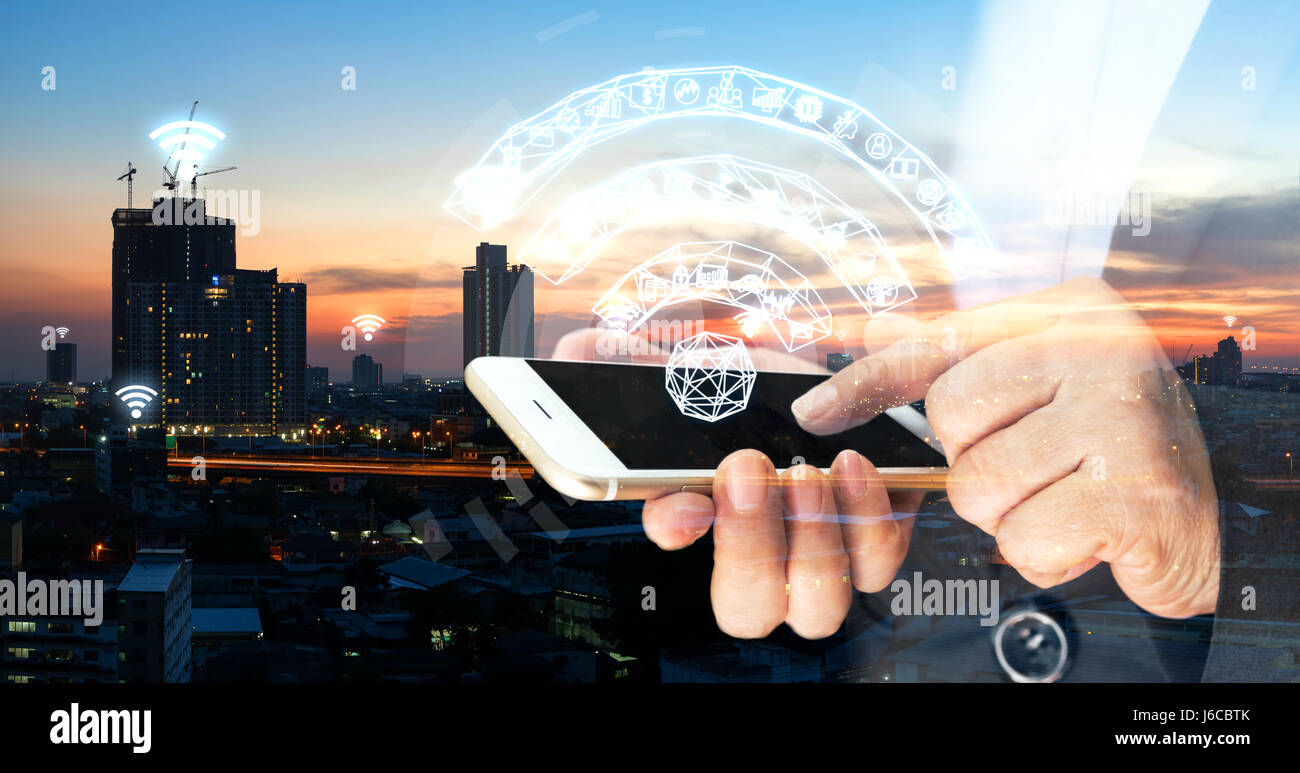 Internet of things , iot , smart home , smart city and network connect concept. Human hand holding white phone and iot icon with city sunset view back Stock Photo