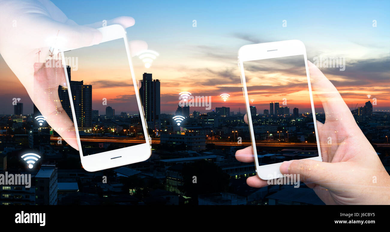 Internet of things , iot , smart home , smart city and network connect concept. Hands holding white phones and wifi icon with city sunset view. Stock Photo