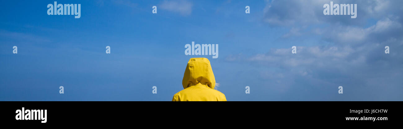 An extreme wide angle image of the rear view of a woman in a bright yellow hooded jacket under an expansive blue sky. Stock Photo