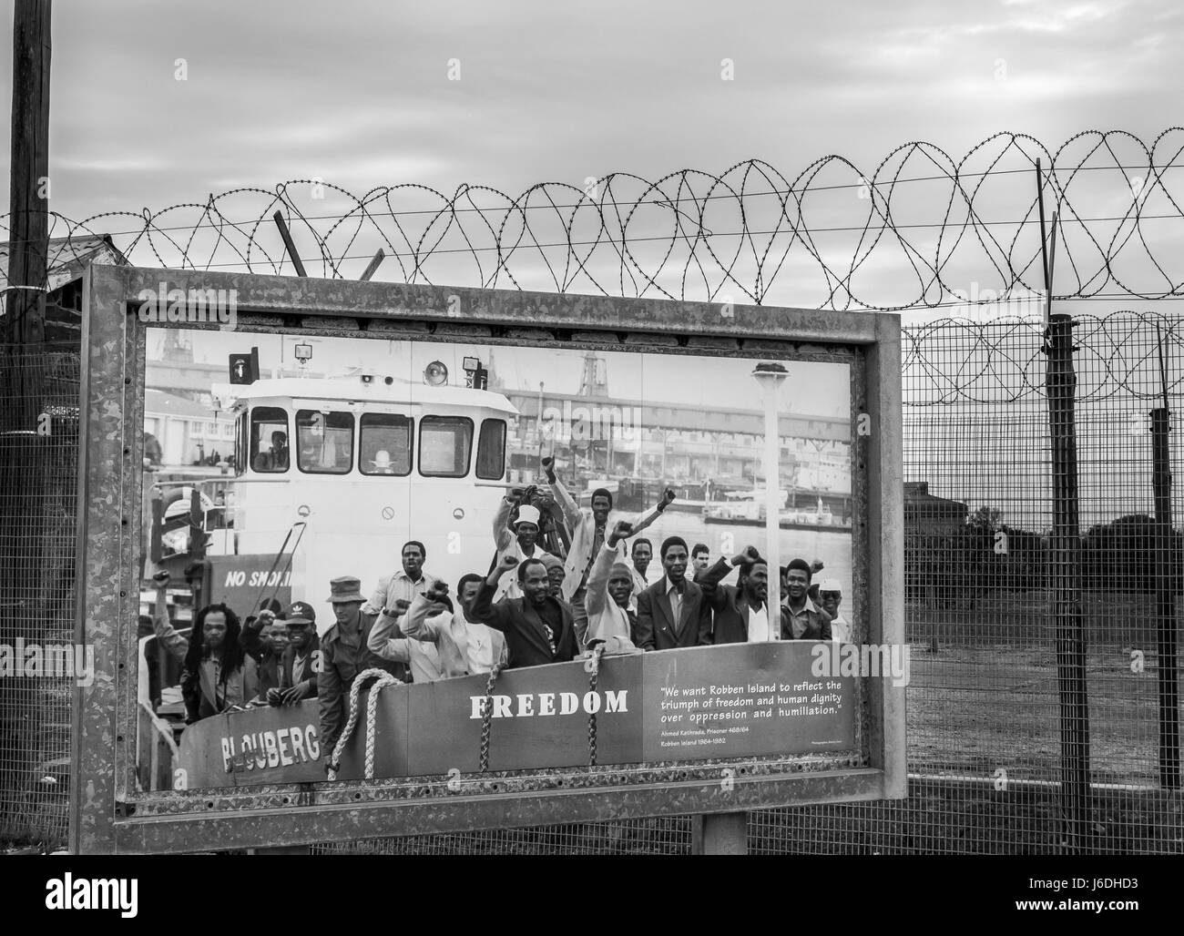 Monochrome photo of poster of last political prisoners to be freed from Robben Island near barbed wire ringed prison entrance, Cape Town, South Africa Stock Photo
