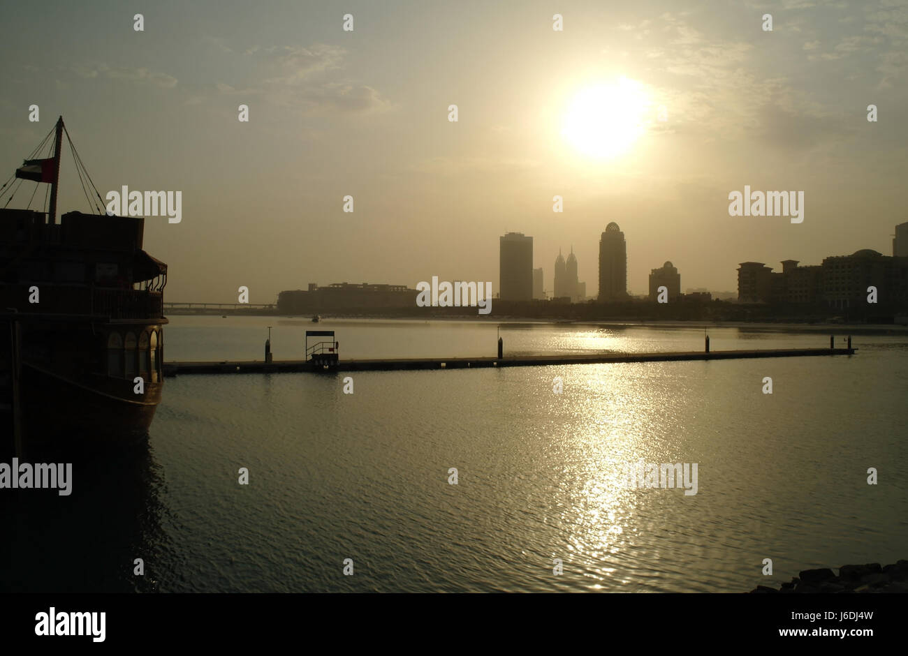View from Al Sufouh Road by stern of Aprons and Hammers Dhow across Mina Seyahi Beach Resort to sun rising above silhouette skyscrapers, Dubai, UAE Stock Photo