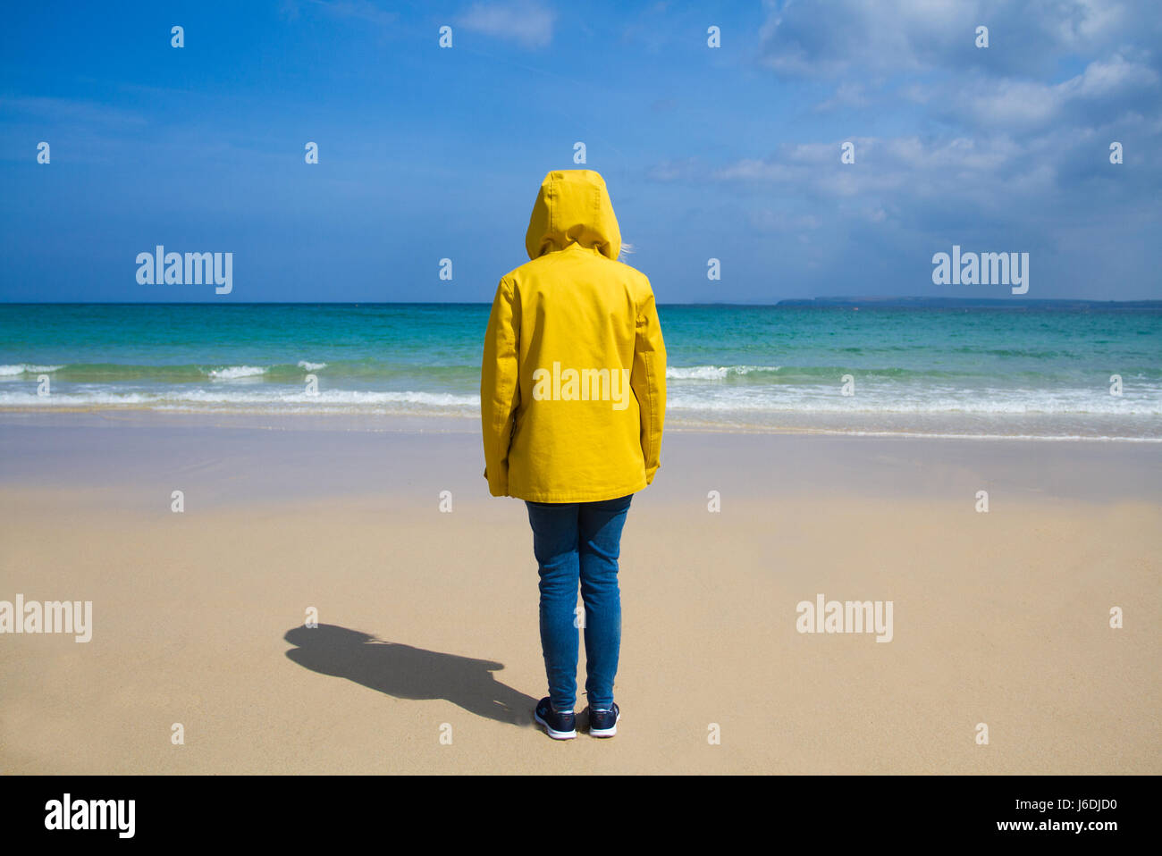 A single woman wearing a bright yellow, hooded jacket whilst standing on a deserted, sandy beach on a tropical paradise island. Stock Photo