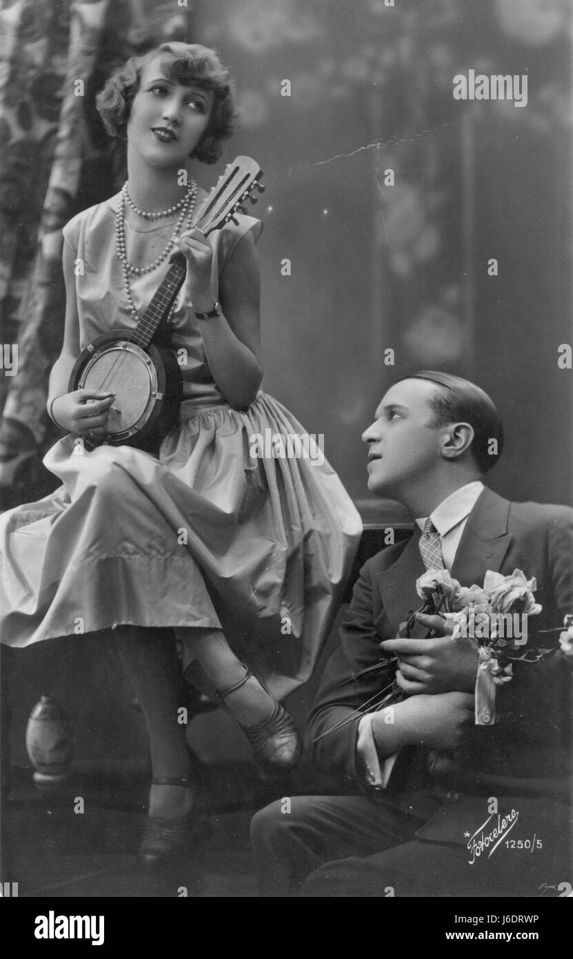 Woman playing Banjo and singing to a man who is gazing fondly upward at the musician lady, as he clutches a bouquet of  flowers (roses?).  She wears 3 strings of Pearls, and he has French cuff links in his shirt.  Probably late 1920s.   To see my related vintage images - in Search:  Prestor  vintage  music  womanflo Stock Photo