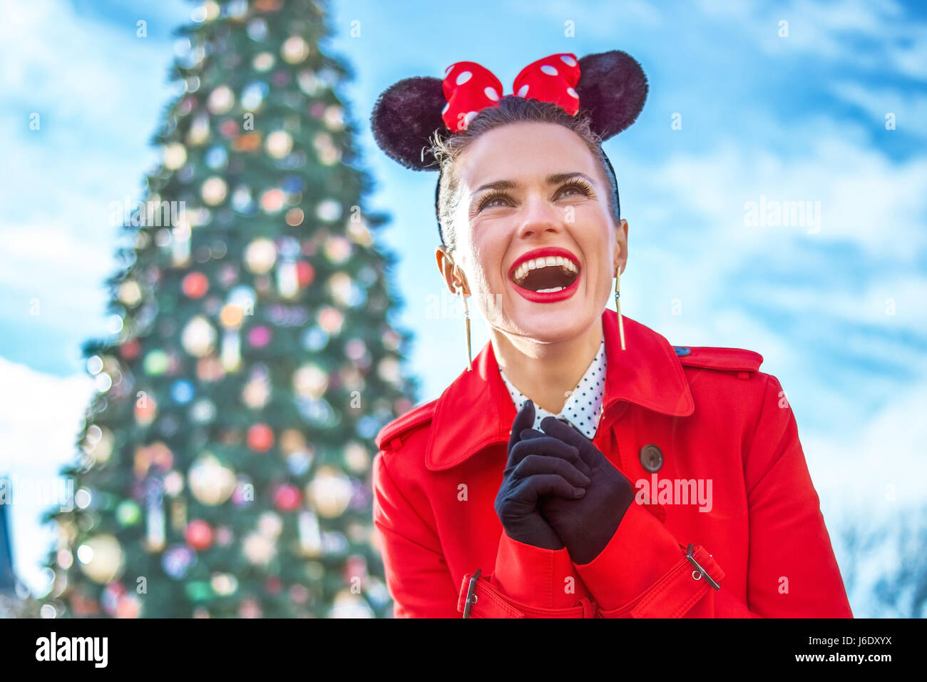 DISNEYLAND, FRANCE - DECEMBER, 8, 2016: smiling trendy woman in red trench coat in the front of big Christmas tree looking into the distance Stock Photo