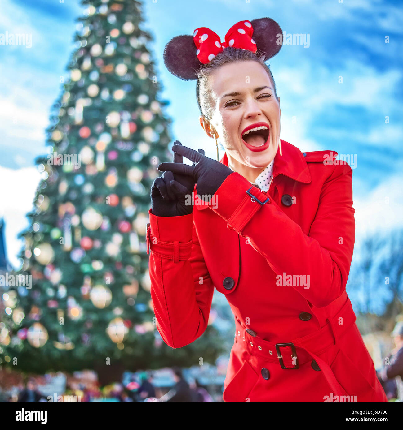 DISNEYLAND, FRANCE - DECEMBER, 8, 2016: smiling young woman in red trench coat in the front of big Christmas tree showing hashtag gesture Stock Photo