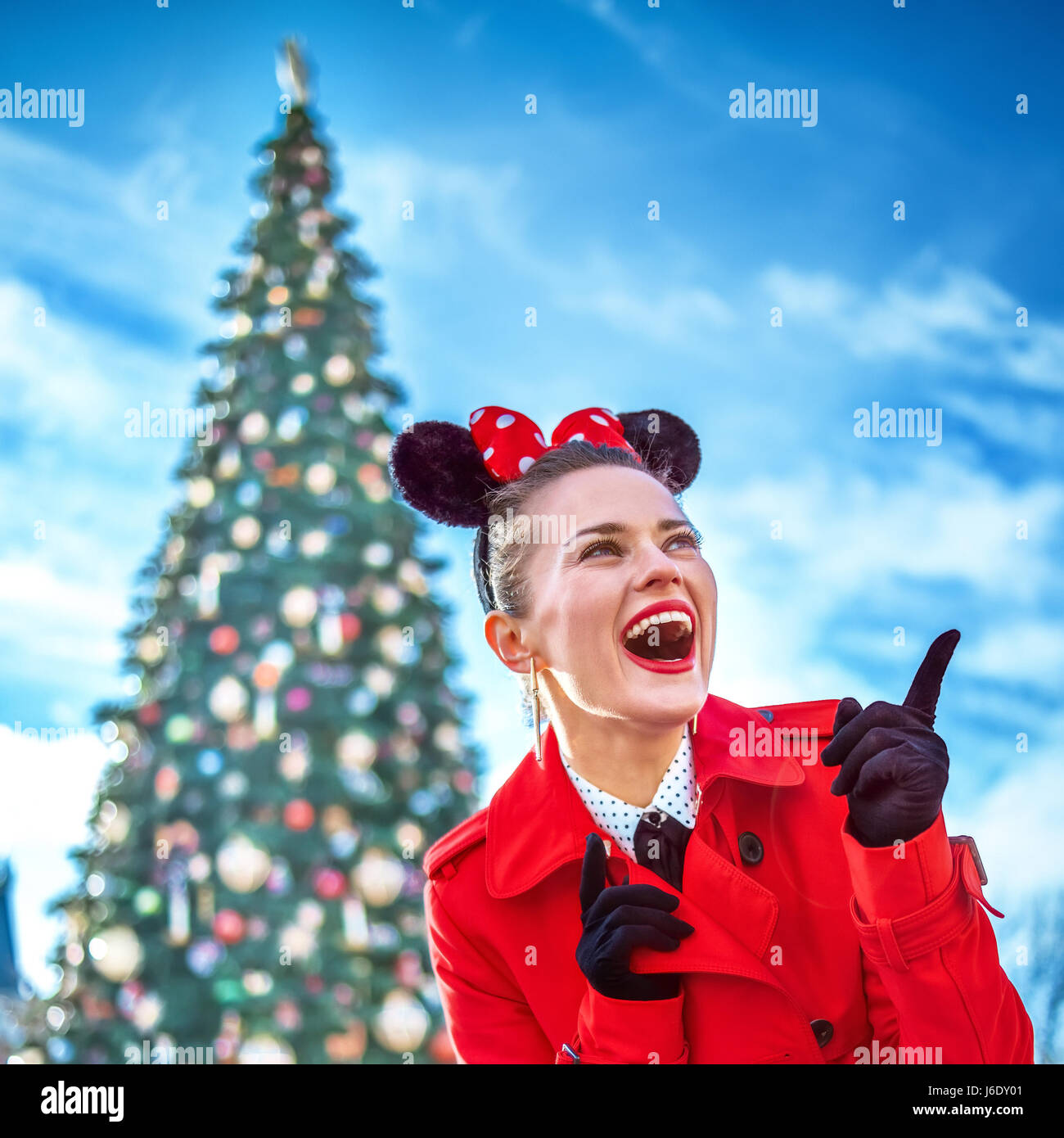 DISNEYLAND, FRANCE - DECEMBER, 8, 2016: happy modern woman in red trench coat in the front of big Christmas tree pointing at something Stock Photo
