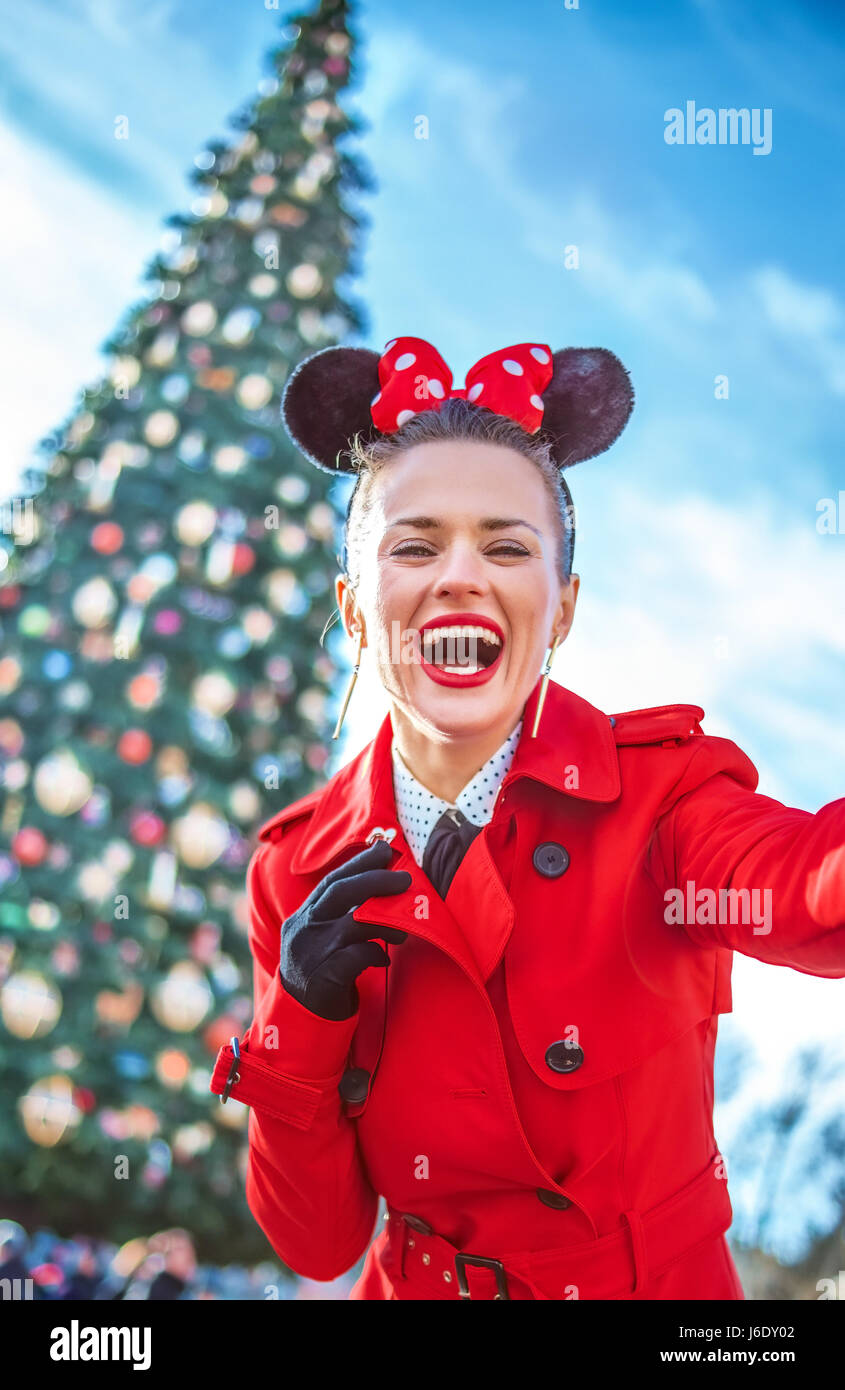 DISNEYLAND, FRANCE - DECEMBER, 8, 2016: happy stylish woman in red trench coat in the front of big Christmas tree taking selfie Stock Photo