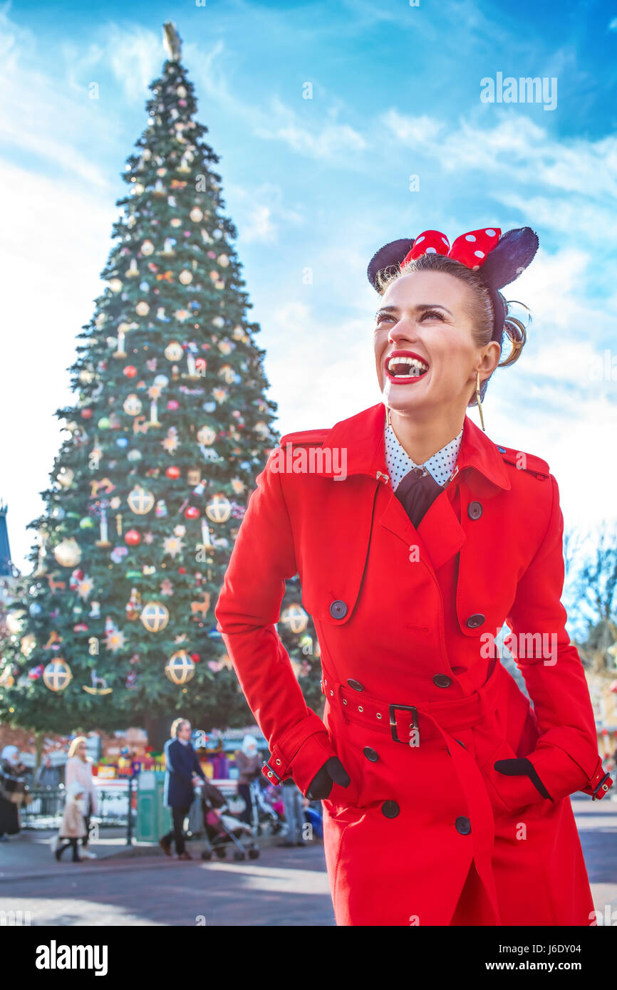 DISNEYLAND, FRANCE - DECEMBER, 8, 2016: smiling elegant woman in red trench coat in the front of big Christmas tree looking into the distance Stock Photo