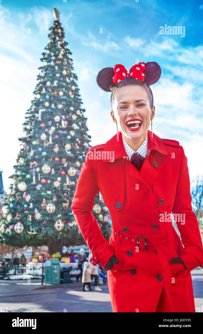 DISNEYLAND, FRANCE - DECEMBER, 8, 2016: Portrait of happy stylish woman in red trench coat standing in the front of big Christmas tree Stock Photo